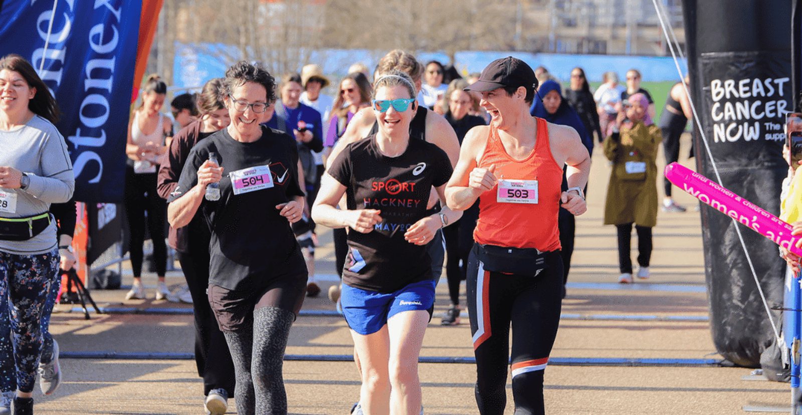 Three women smiling and running together near a race finish line, surrounded by other participants and spectators. A "Breast Cancer Now" banner is visible in the background.