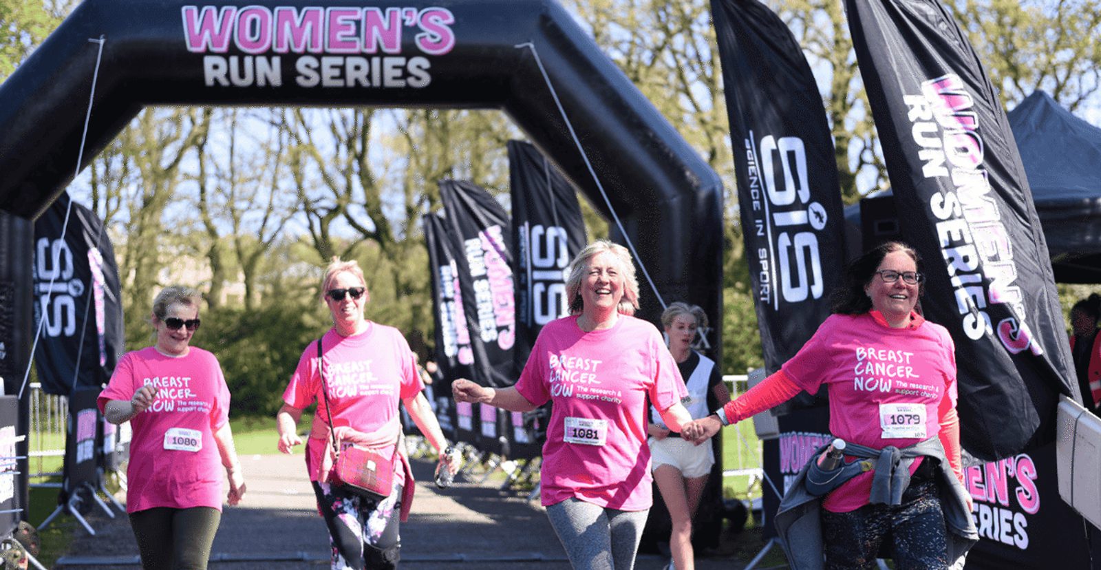 Five women wearing pink “Breast Cancer Now” shirts cross the finish line together at a Women’s Run Series event. They are smiling and holding hands, surrounded by banners and trees.