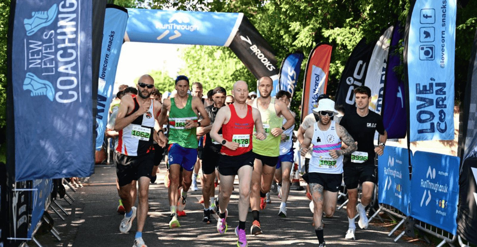A group of runners begins a race under a blue inflatable arch, surrounded by banners and flags, on a sunny day. Some runners wear sunglasses and hats, and trees line the path in the background.