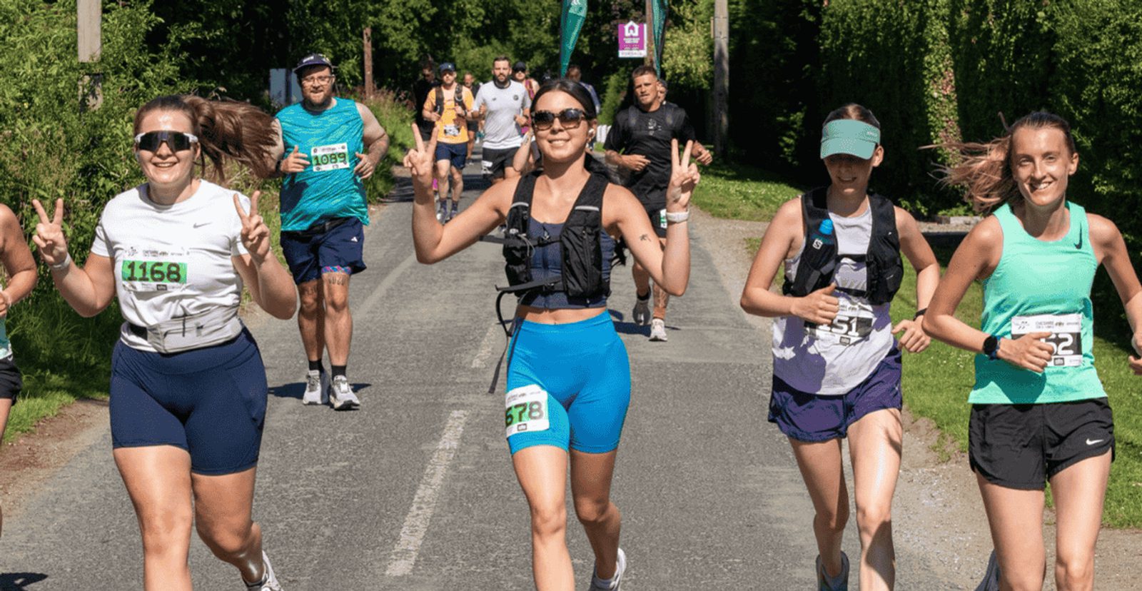 A group of runners, both men and women, smile and make peace signs as they participate in a race on a sunny day, surrounded by greenery. Race bibs are visible on their shirts.