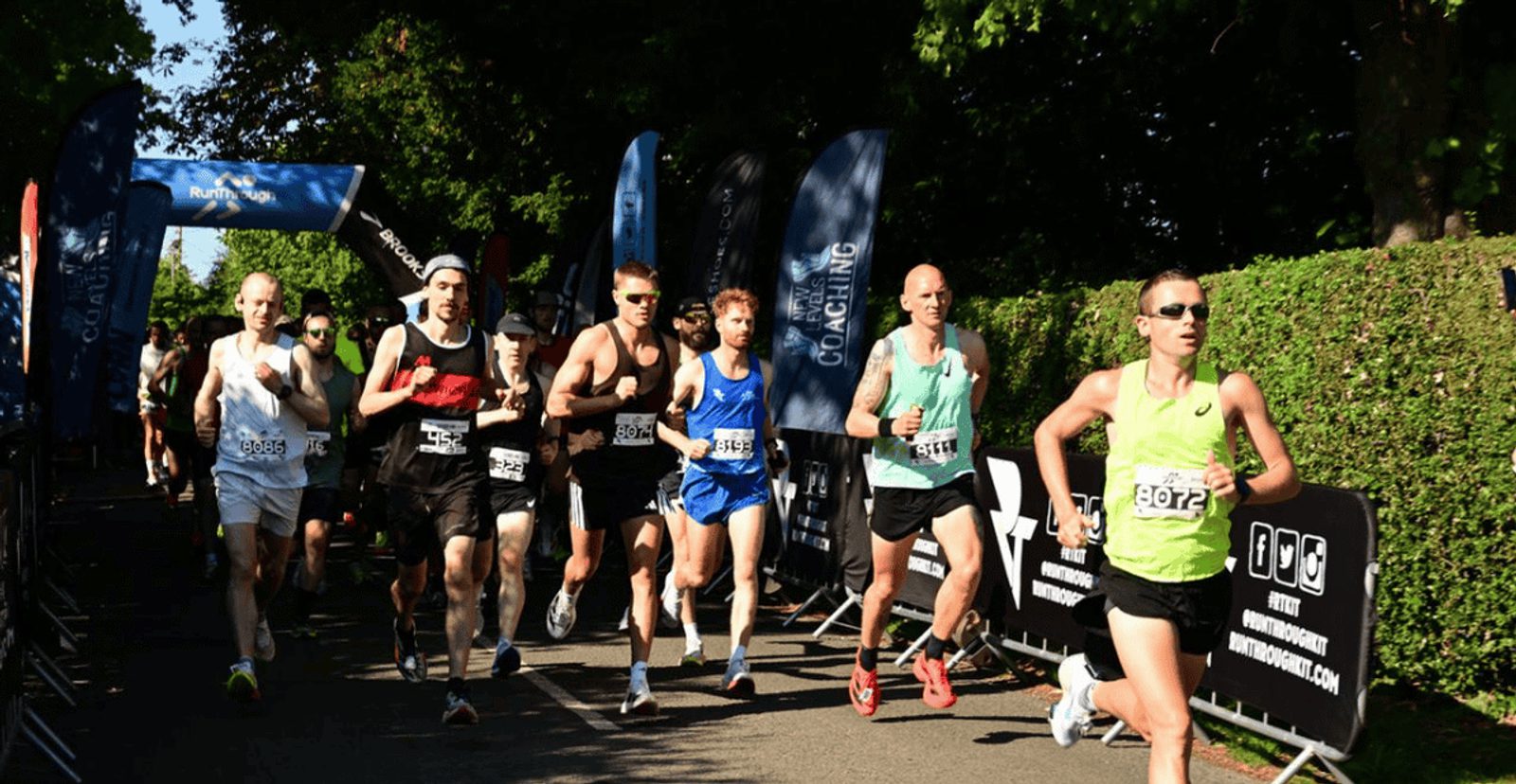 A group of runners in athletic gear participate in a race on a sunny day, passing under a blue race banner, with greenery and event signs along the route.