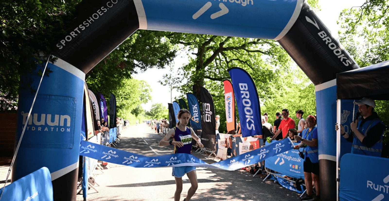 A runner crosses the finish line in first place at a race, breaking the blue tape under an archway marked with "Brooks" and surrounded by cheering spectators and event banners.