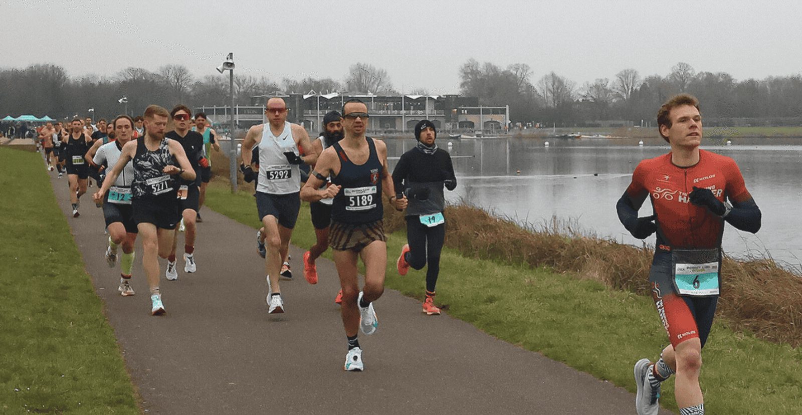A group of athletes running on a paved path beside a lake during a race; the weather is overcast, and the runners wear numbered bibs and athletic gear. Trees and a building are visible in the background.