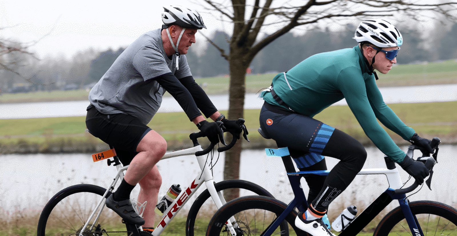 Two cyclists ride on a path beside a body of water. One wears a gray shirt and black shorts, the other wears a teal jacket and blue leggings. Both wear helmets and sunglasses, with trees and grass visible in the background.