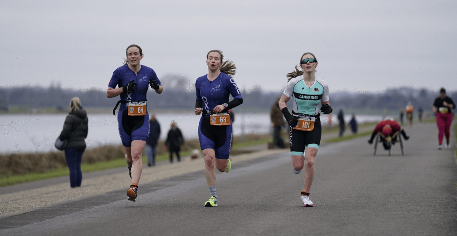 Three women wearing race bibs run on a paved path near water, with other runners and a person in a wheelchair visible in the background on an overcast day.