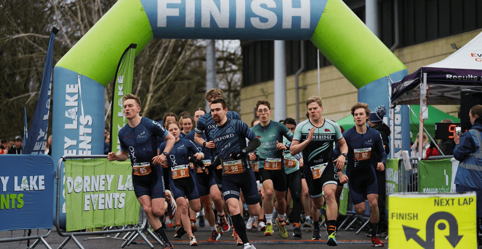 A group of athletes in racing gear start running from a starting line under a large inflatable arch labeled "FINISH" at a competitive outdoor event. Spectators and event banners are visible in the background.