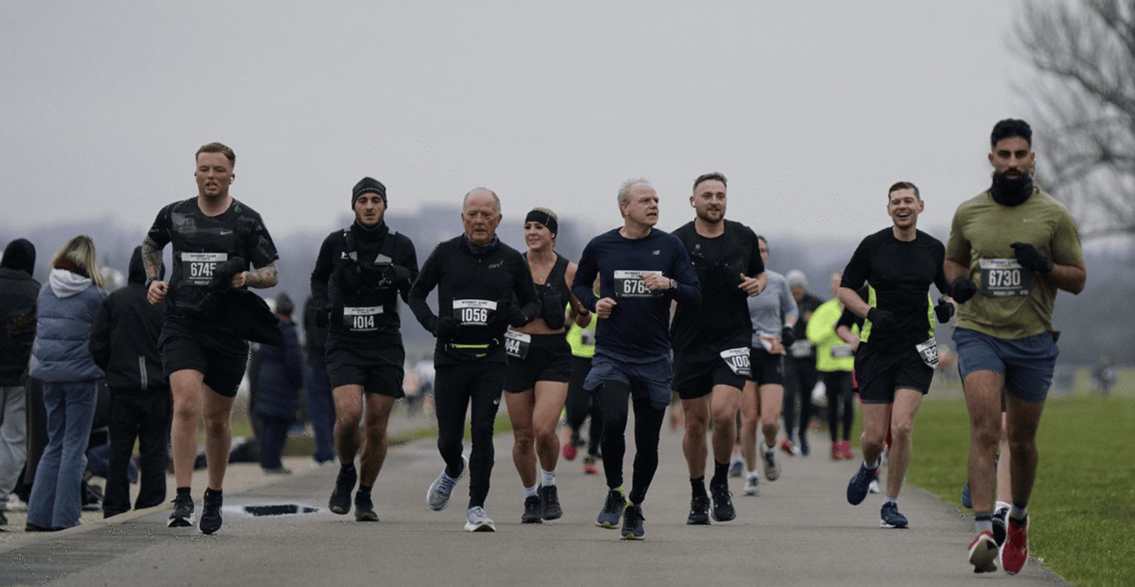 A group of runners, dressed in athletic gear and race bibs, run together on a paved path during a race. The background is cloudy, with spectators and more runners visible.