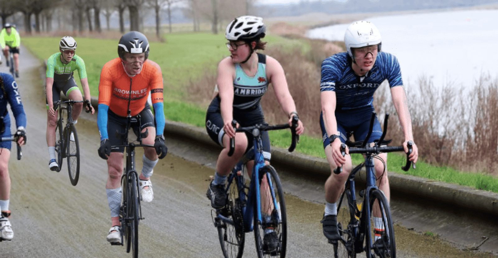 Four cyclists ride along a riverside path, two in the foreground wearing helmets and kits labeled "Cambridge" and "Oxford," and two more cyclists following behind them on the same road. Trees line the path.