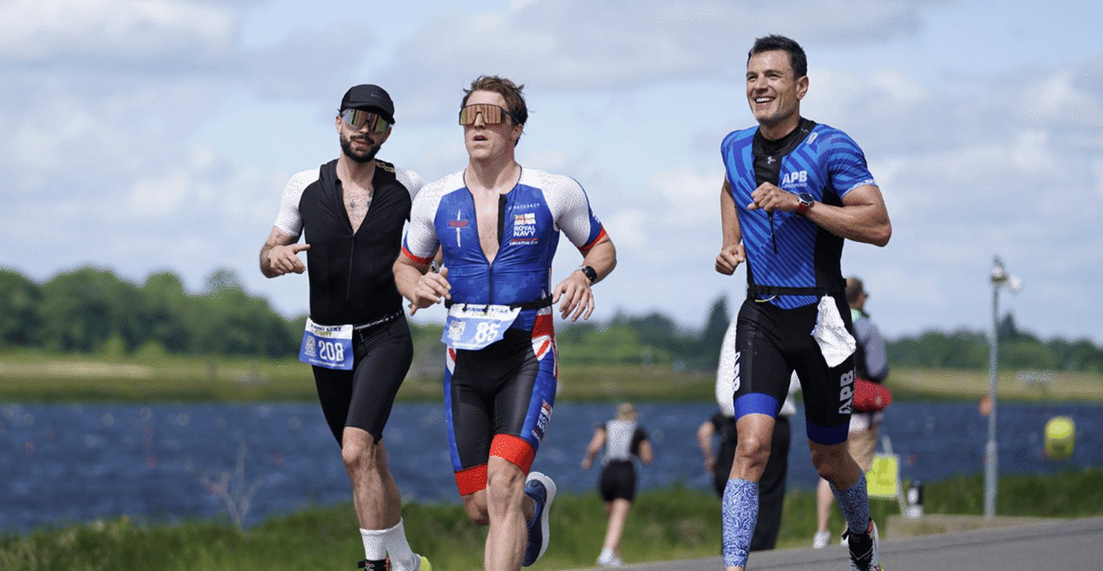 Three male athletes in sportswear run on a road near a body of water during a race, with trees and blue sky in the background. The runners are focused, and one is smiling.