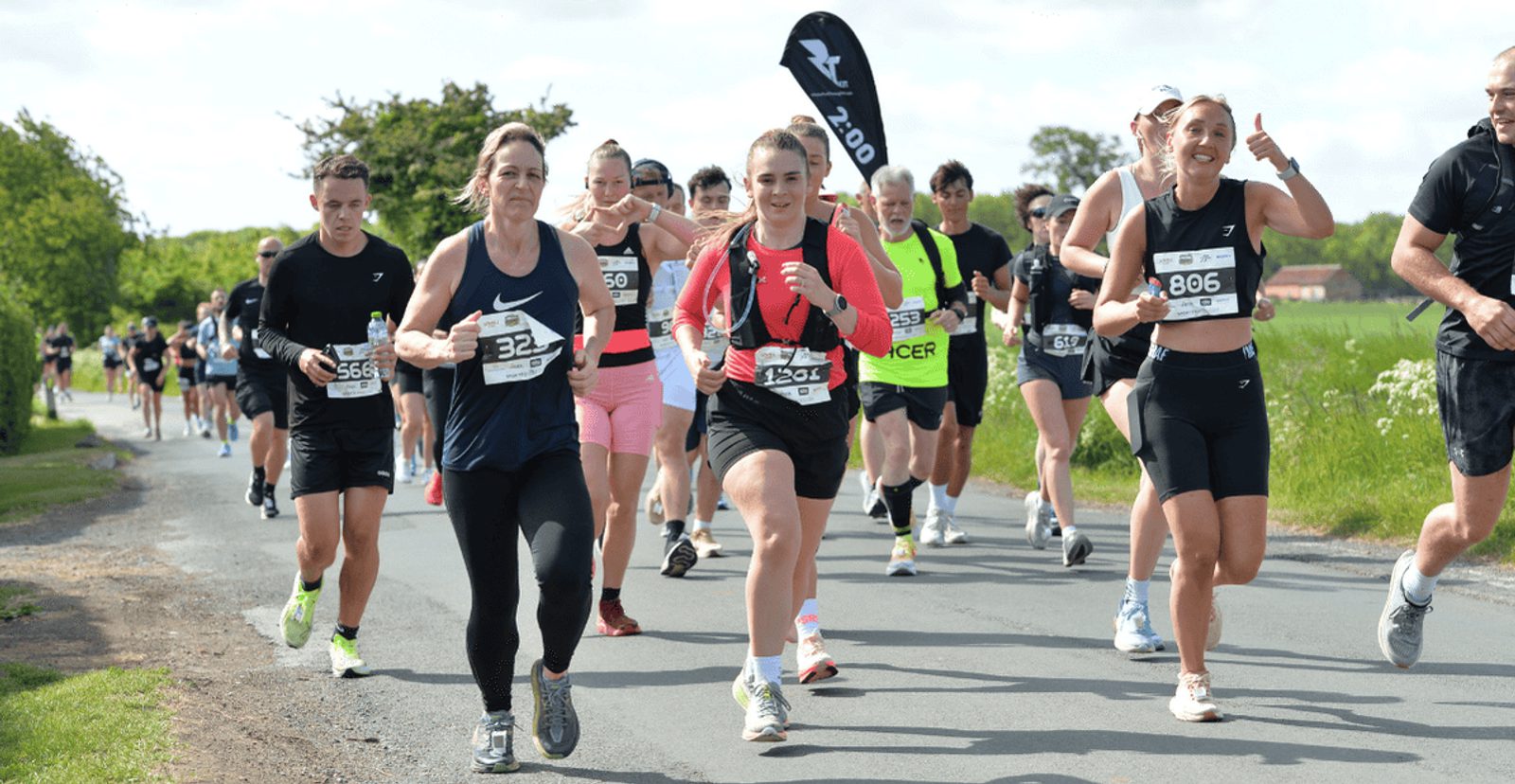 A group of runners of various ages participate in a road race on a sunny day, wearing bib numbers and athletic gear. Some are smiling, and one runner gives a thumbs up. Trees and grass line the roadside.