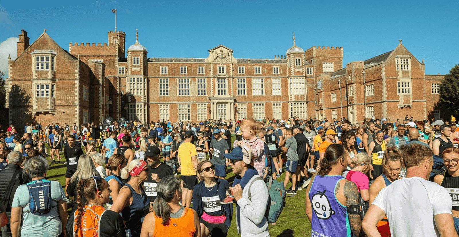 A large crowd of runners and spectators gathers on a sunny day in front of a grand historic brick building with many windows, preparing for a race. Some people wear running gear and numbered bibs, while others watch or socialize.