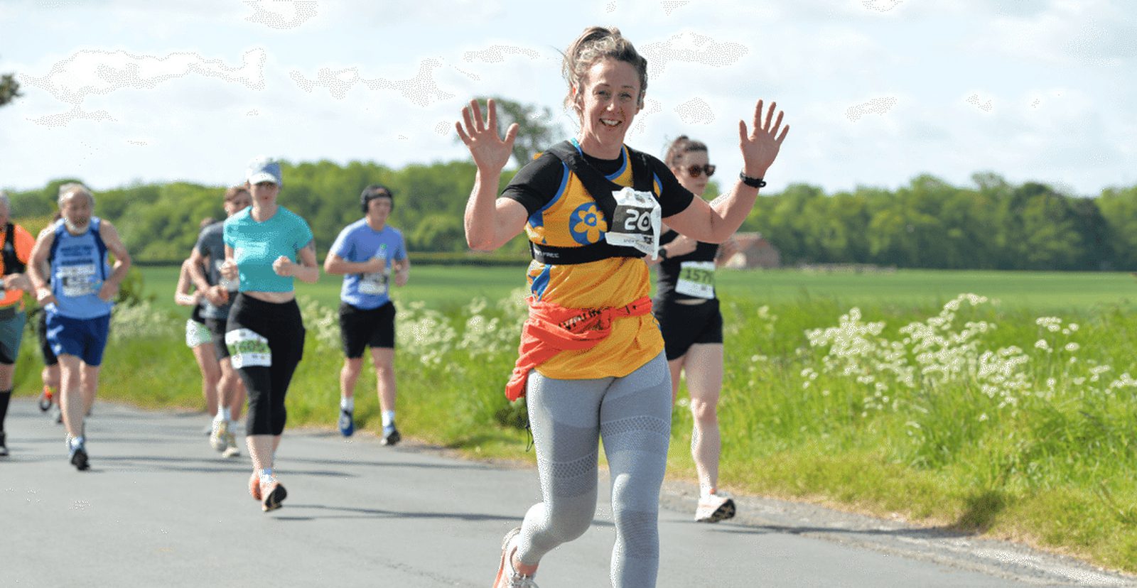 A woman runner in a yellow and black shirt smiles and raises her hands while running on a road with other runners behind her. The scene is outdoors on a sunny day with green fields in the background.