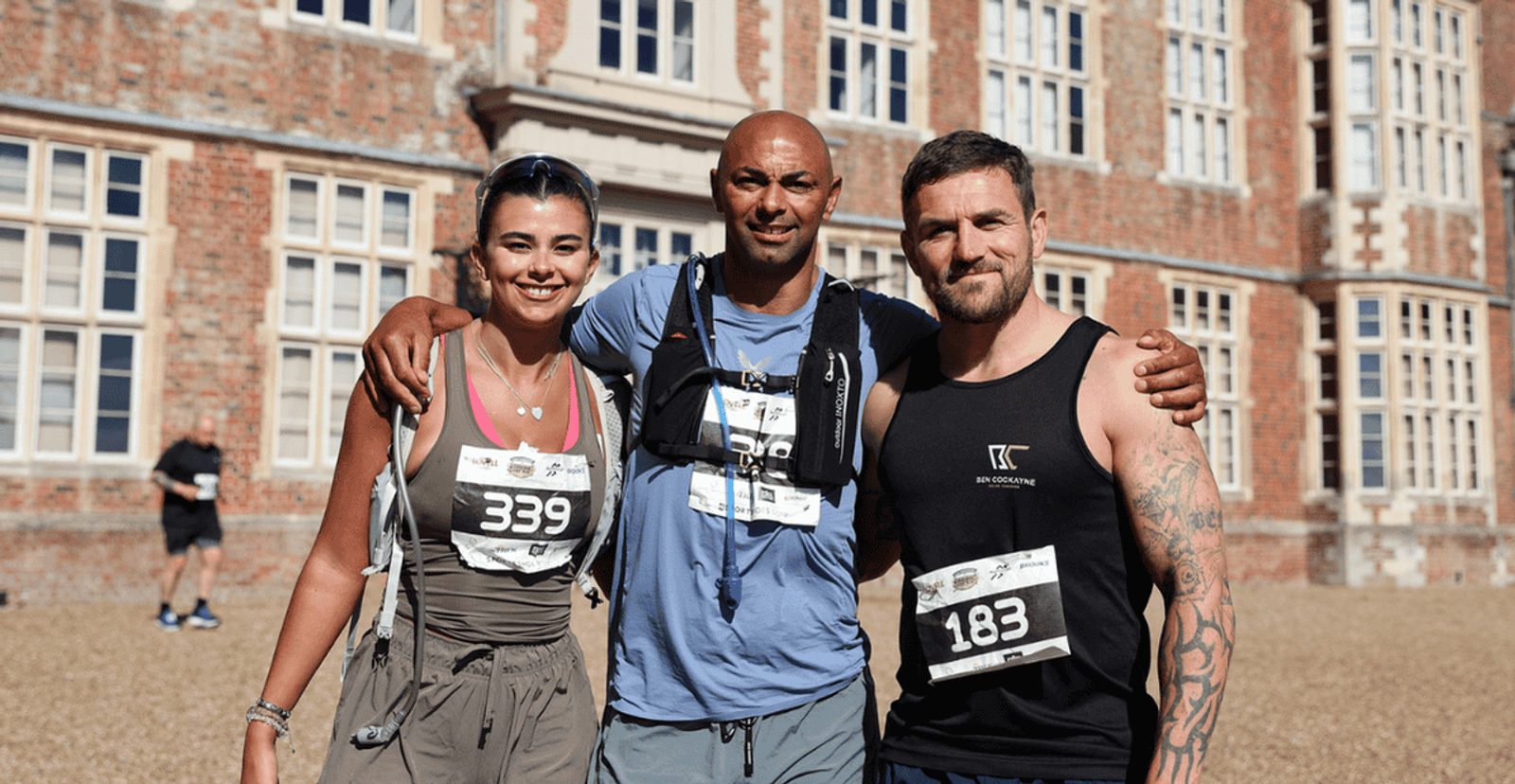 Three runners wearing numbered race bibs pose together and smile in front of a large, historic brick building with many windows on a sunny day.