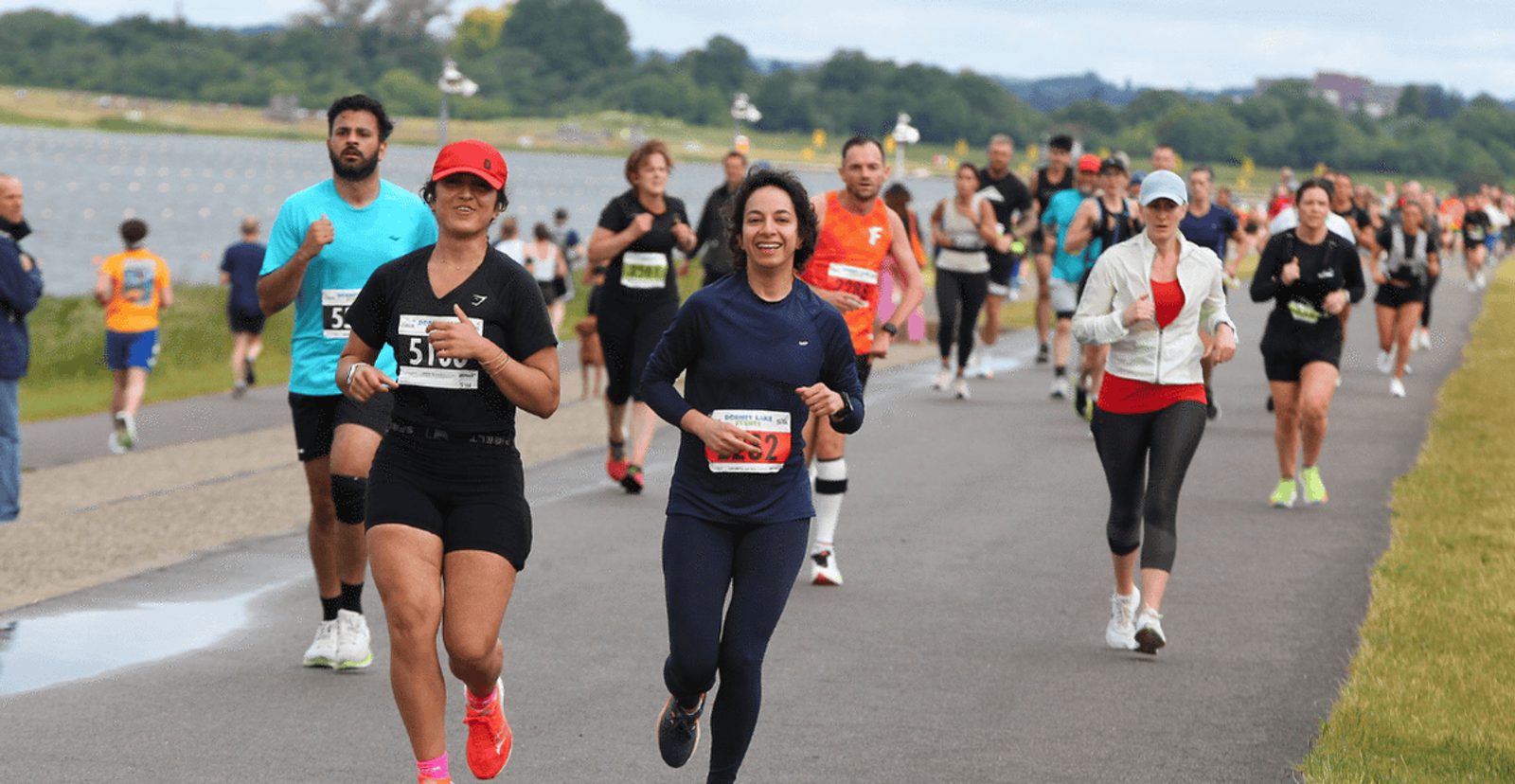 A group of runners, both men and women, compete in a race along a paved path outdoors, with some smiling and wearing numbered bibs. Greenery and more runners are visible in the background.