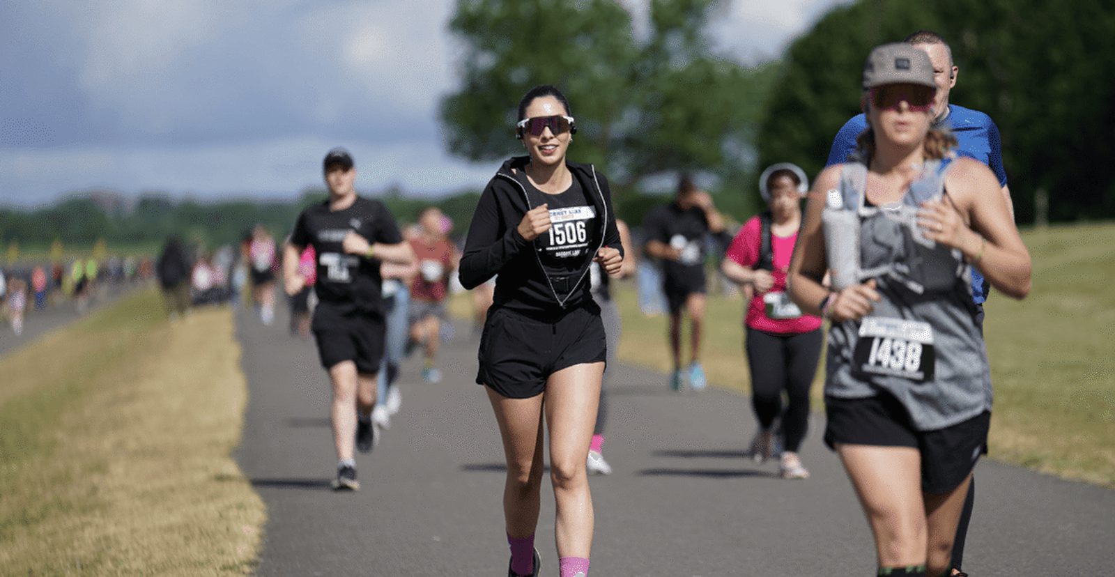 A group of people of various ages running outdoors on a paved path during a race, with race bibs on their clothing and trees in the background. The weather appears sunny.