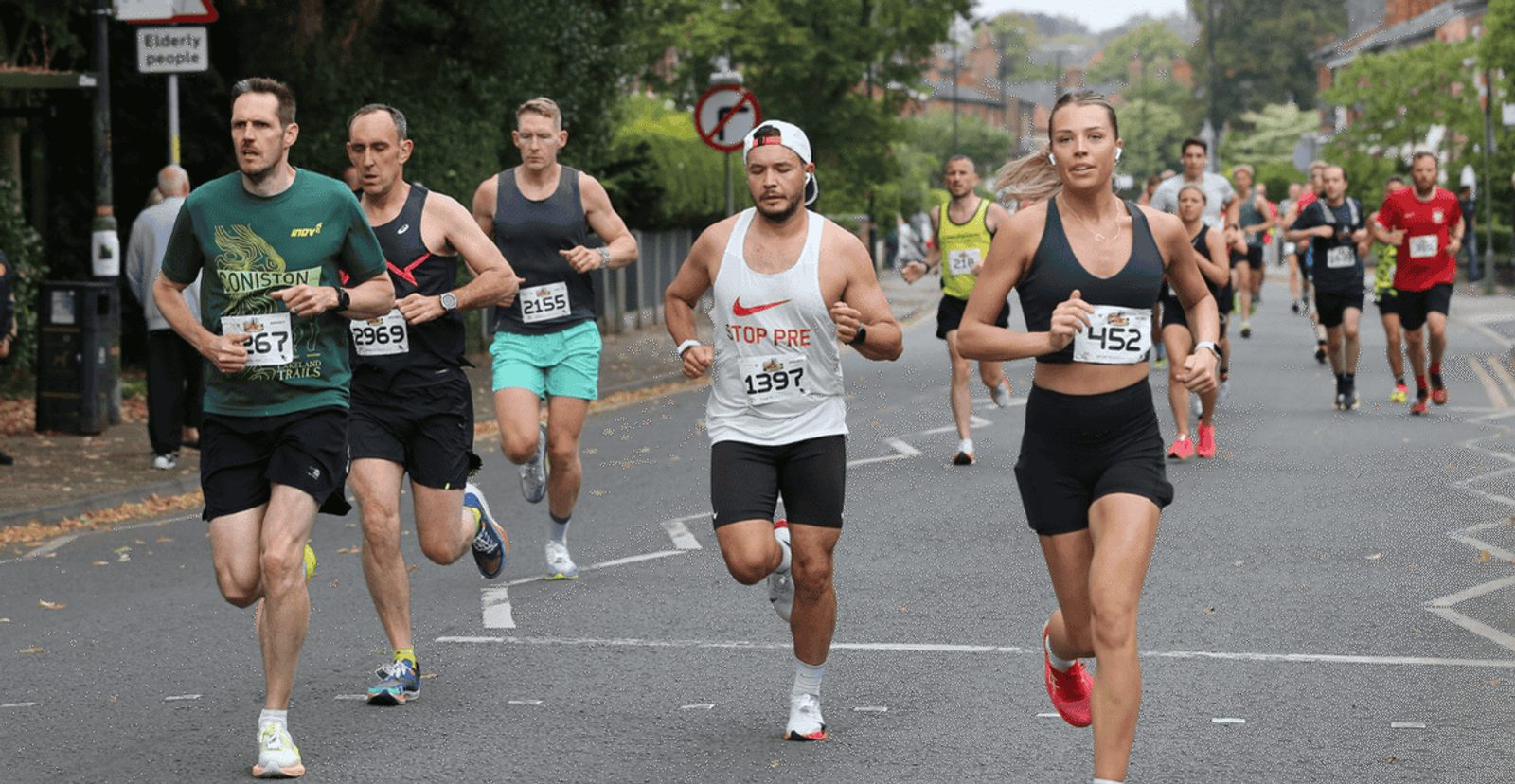 A group of runners, both men and women, participating in a road race, wearing numbered bibs and athletic gear, run along a tree-lined street with more participants visible in the background.