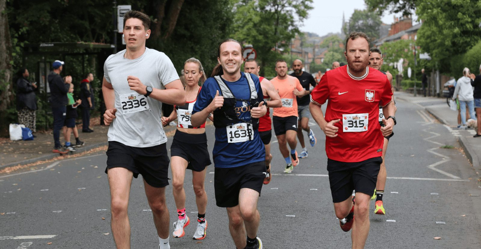 A group of runners, each wearing a numbered bib, participate in a road race on a city street lined with trees and spectators. The runners look focused and determined as they move forward.