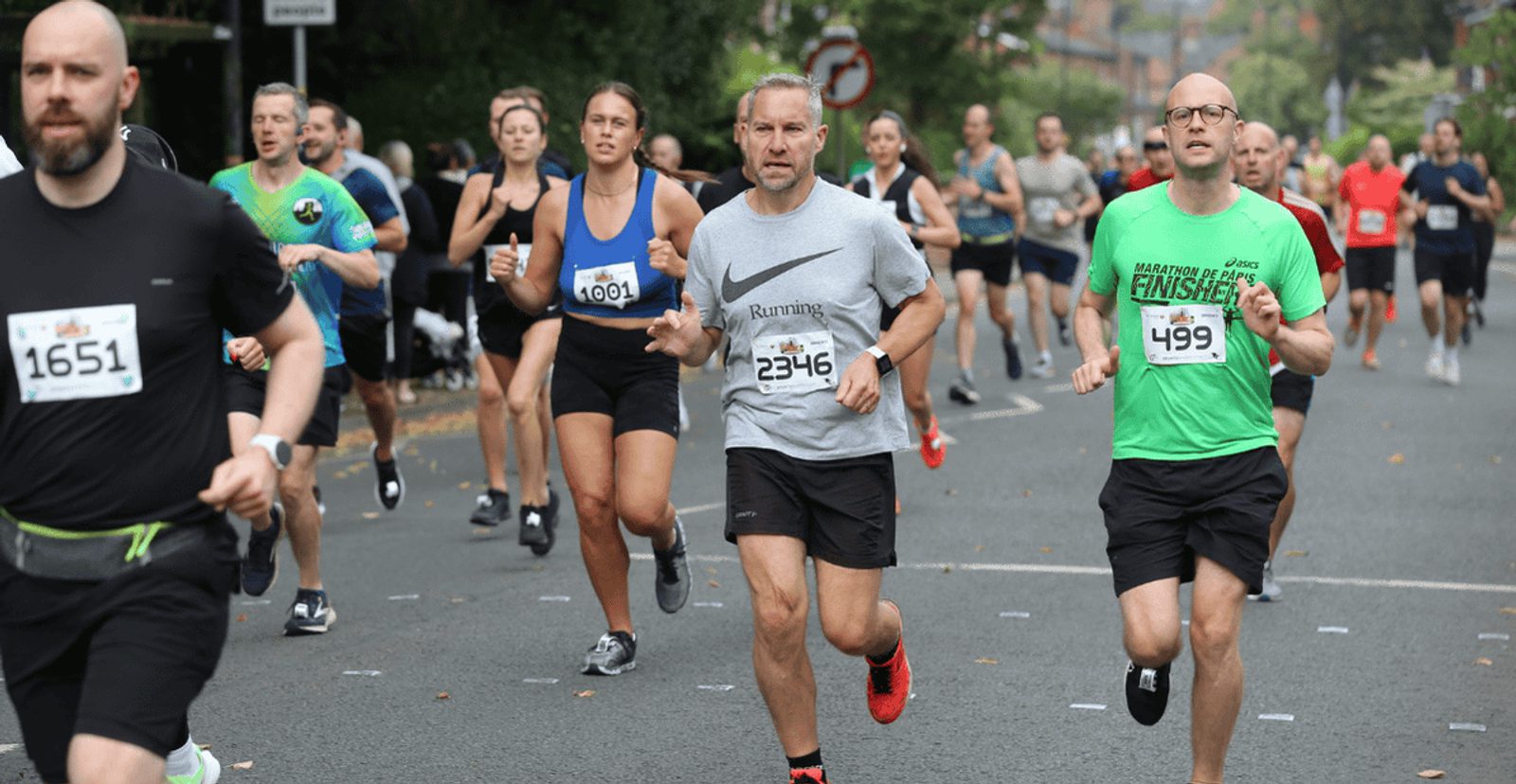 Runners participate in a road race, wearing numbered bibs. Some look focused, others relaxed. Trees and spectators line the road in the background on a cloudy day.