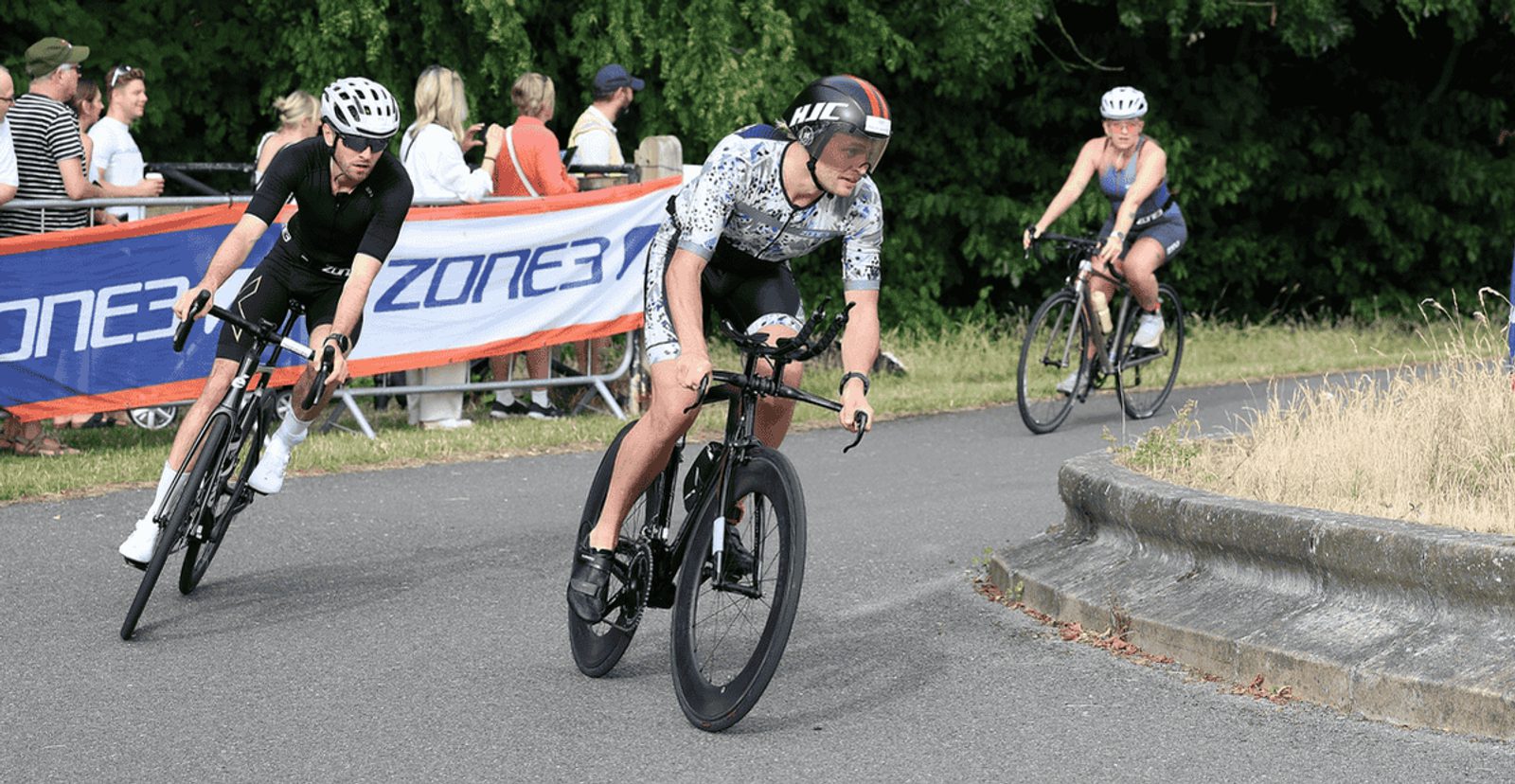 Three cyclists in helmets race around a curve on a paved road, with spectators standing behind a banner in the background and greenery surrounding the scene.