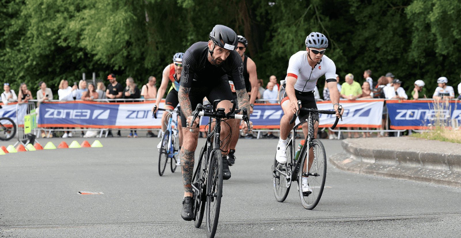Cyclists compete in a road race, riding closely together on a paved course lined with barriers and watched by a crowd of spectators in the background.