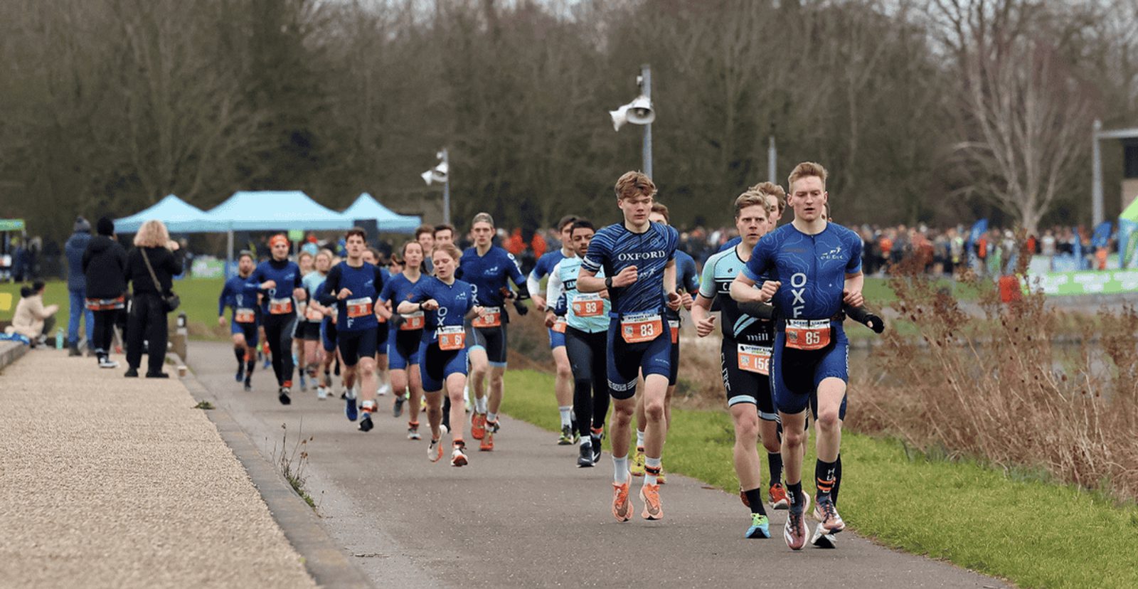A group of athletes in numbered bibs run along a paved path during a race, with trees and spectators in the background on a cloudy day.