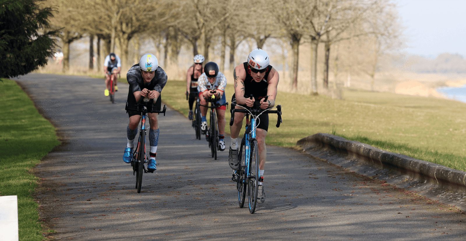 Several cyclists wearing helmets and racing gear ride closely together down a shaded, tree-lined road during a competition or group ride on a sunny day.