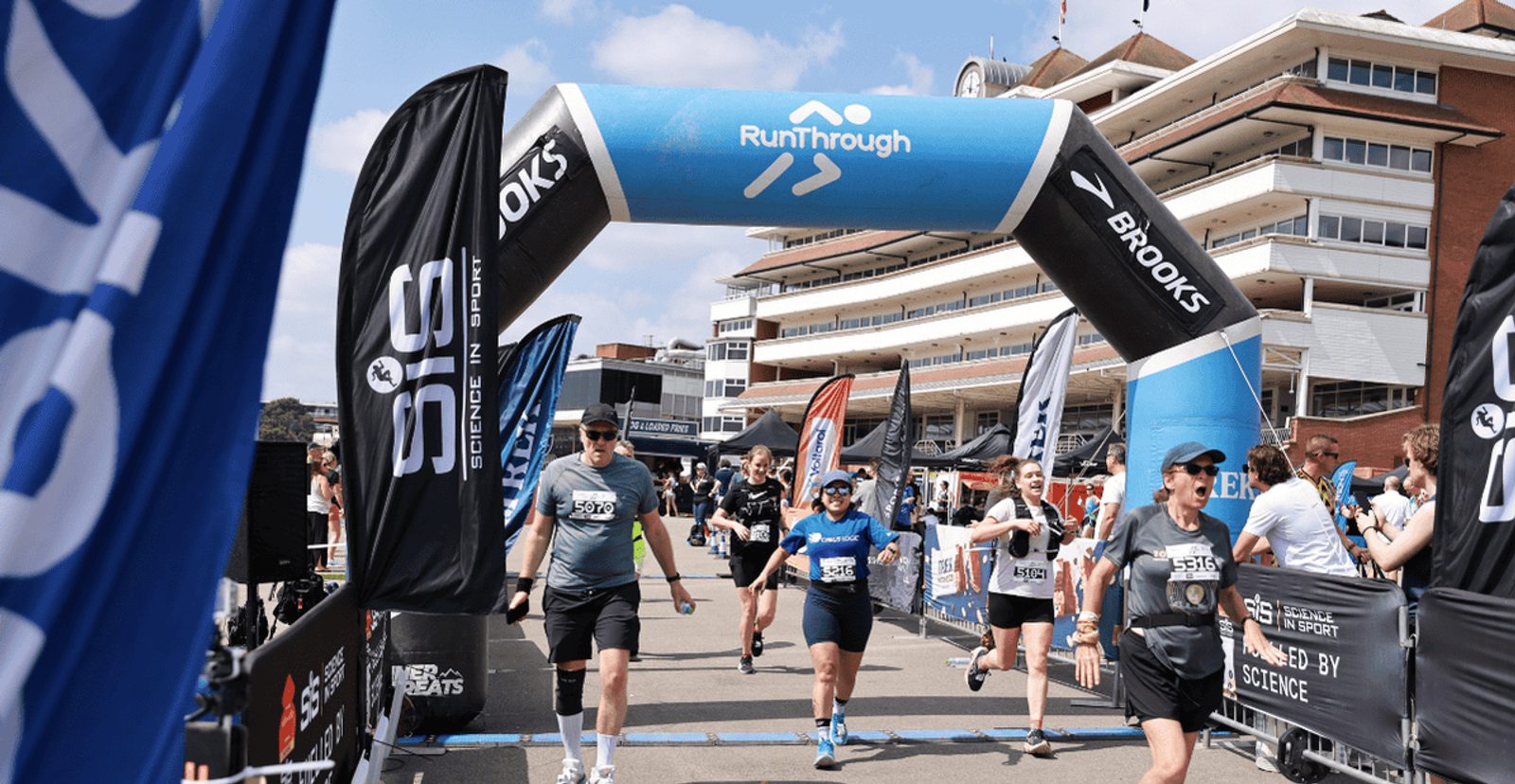 Runners cross the finish line under a "RunThrough" archway at an outdoor event, with flags and banners on both sides and a large building in the background on a sunny day.