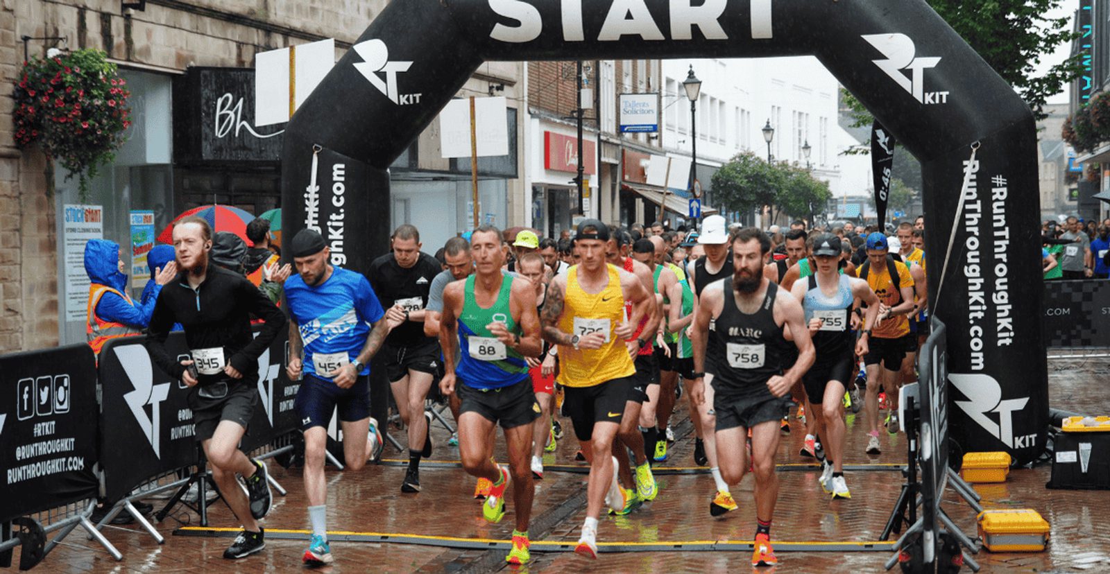 Runners wearing numbered bibs begin a race under a black inflatable "START" arch on a wet city street, surrounded by spectators and race banners.