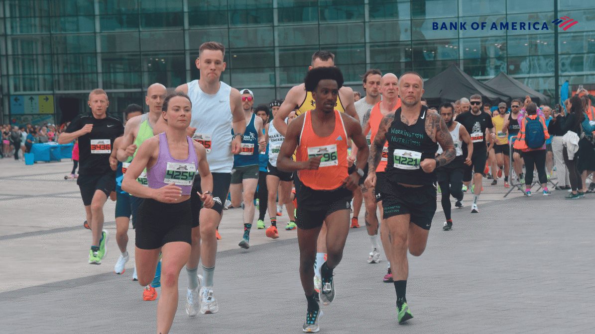 A group of runners, both men and women, competing in a race outside an urban glass building. Some wear numbered bibs and athletic gear. The Bank of America logo appears on the upper right.