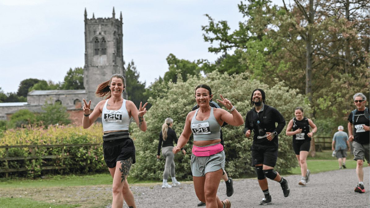 A group of runners smile and make peace signs as they participate in a race on a gravel path, with greenery and a church tower visible in the background.
