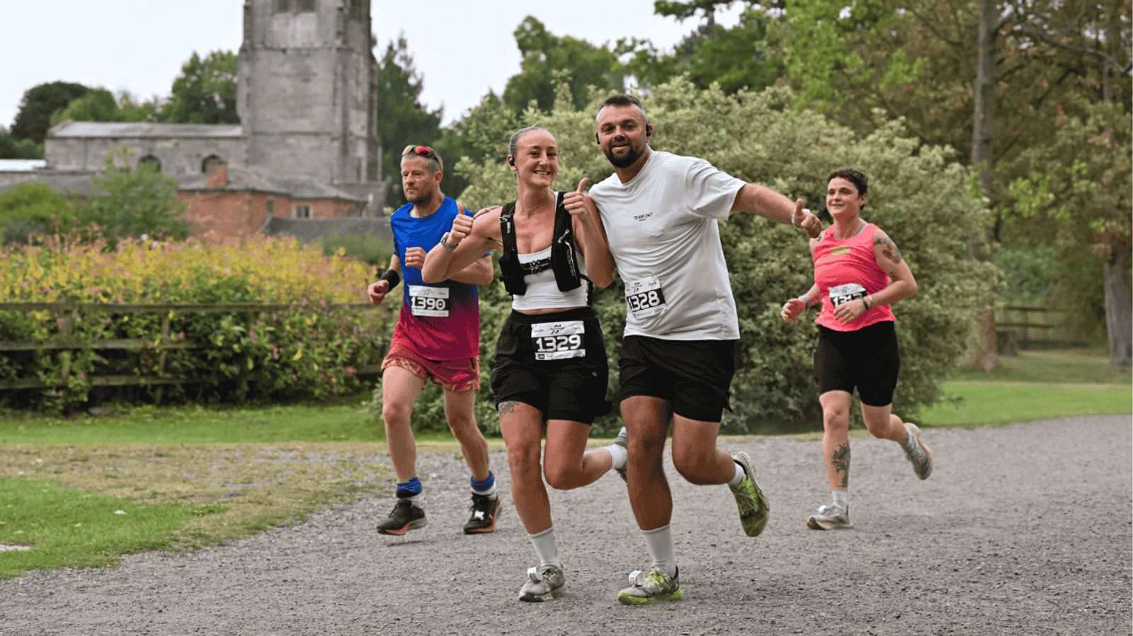 Four runners, wearing numbered race bibs, smile and run on a gravel path through a park. There is greenery and an old stone building in the background. The mood is cheerful and energetic.