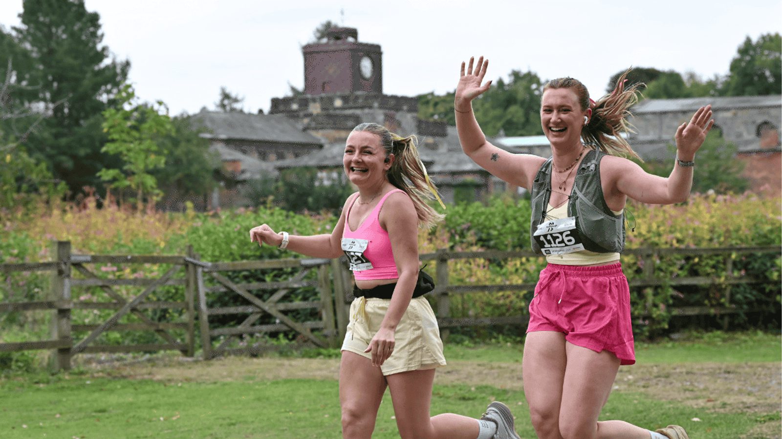 Two women wearing race bibs, athletic tops, and shorts smile and wave as they run through a grassy outdoor area, with trees and a large brick building in the background.