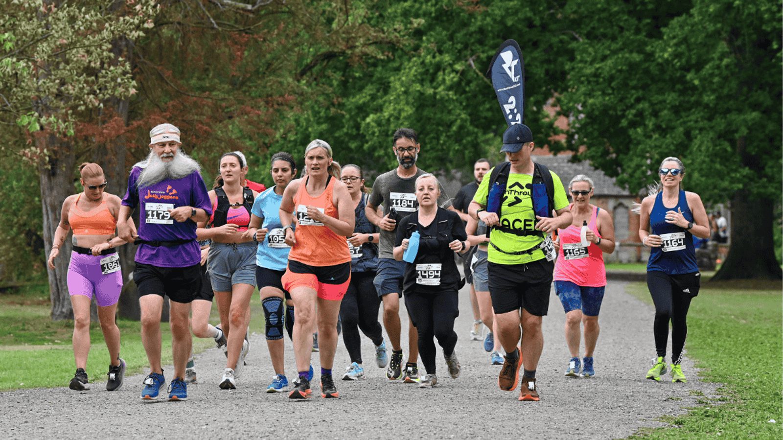 A group of runners of various ages and genders jog together on a park path, wearing race bibs and athletic clothes, surrounded by green trees and grass.