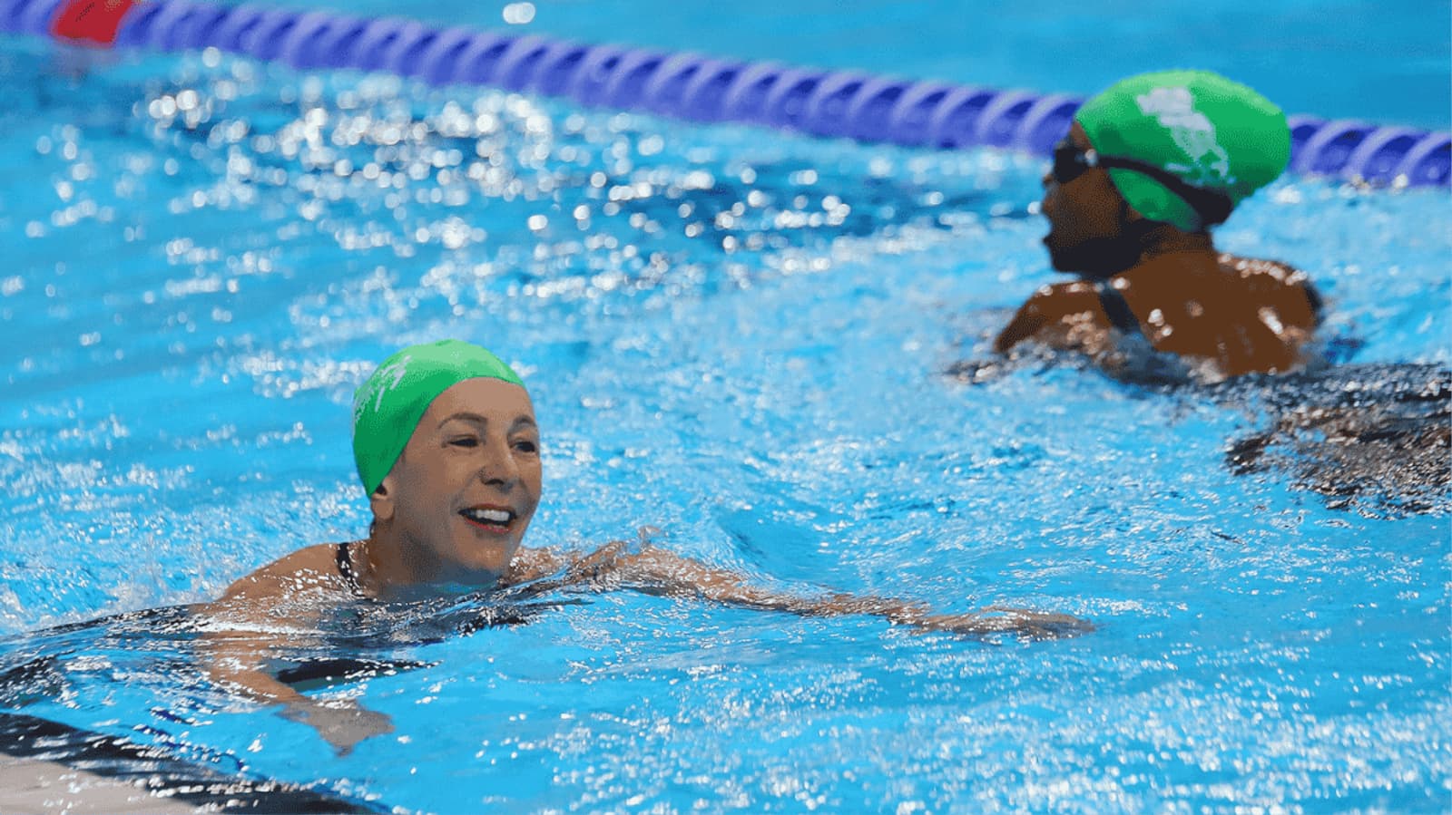 Two swimmers in green caps are in an indoor pool. The swimmer in the foreground is smiling and moving forward, while the other is nearby, facing away. Both are surrounded by blue lane dividers.