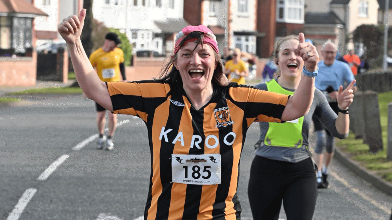 A joyful runner in a black and orange striped shirt, with race number 185, smiles and raises her thumbs up on a residential street. Another runner in a lime green top follows behind, also smiling. Other participants are seen in the background.
