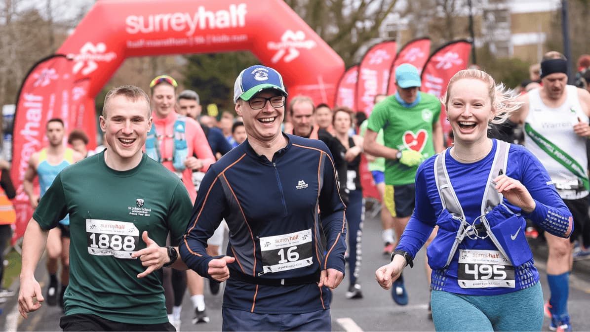 A group of runners, smiling and wearing numbered bibs, participate in the Surrey Half Marathon. They are running in front of a red inflatable arch with event branding. Other runners follow behind them. It's a cloudy day.