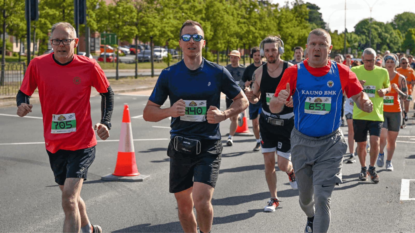 A group of runners participating in a race on a sunny day. They are on a street lined with orange cones, with trees in the background. The runners are wearing race numbers and casual athletic attire, with some in sunglasses.