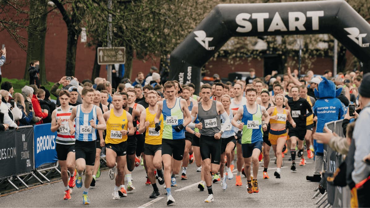 Runners beginning a race at the starting line under a black arch with "START" written on it. They wear numbered bibs and colorful athletic gear. Spectators line the street, and trees and buildings are visible in the background.