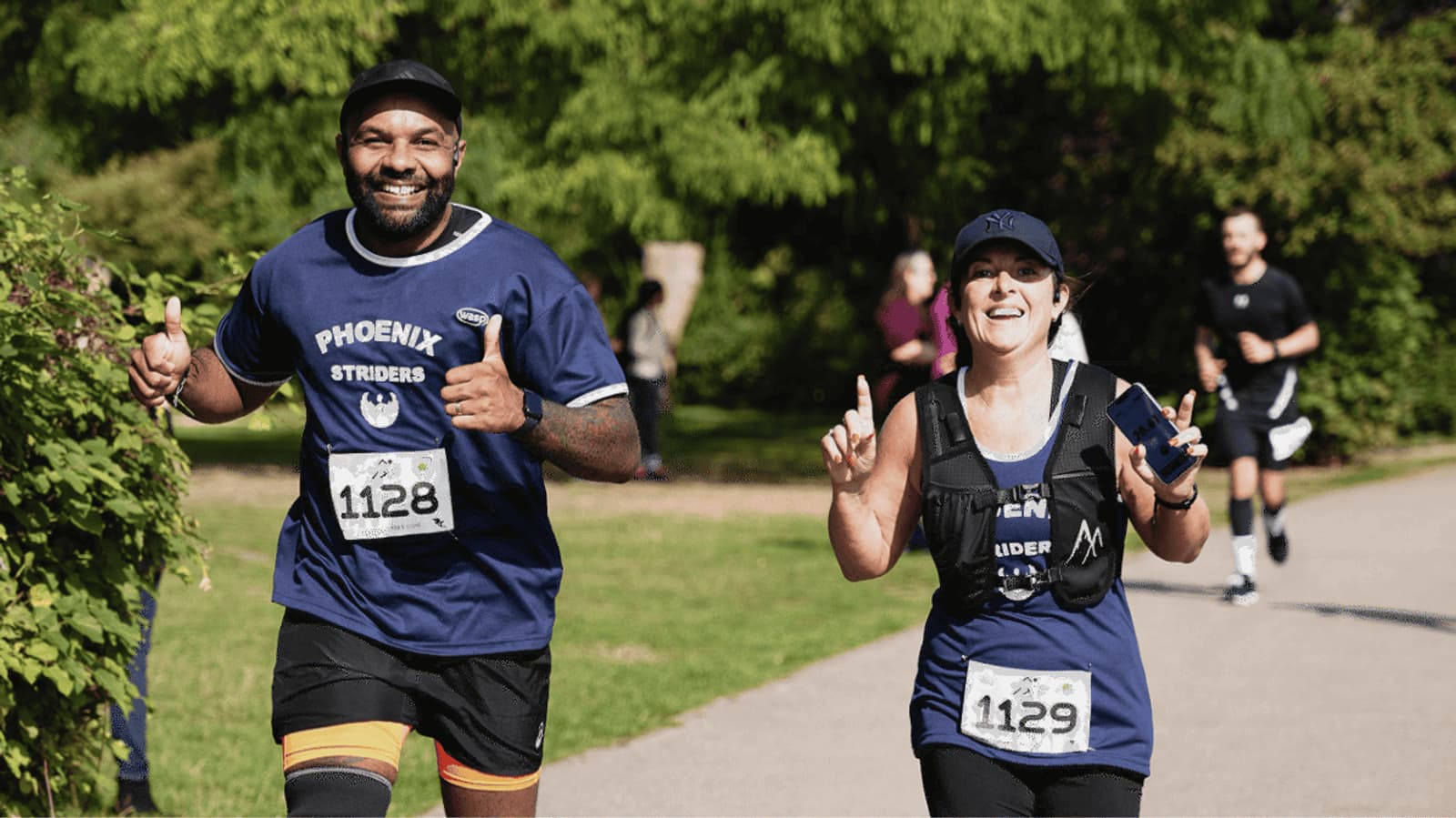 Two runners in blue shirts, smiling and giving thumbs up, participate in a race. The man on the left wears "1128" and the woman on the right, "1129" and a cap. Greenery surrounds the path, and other runners are in the background.