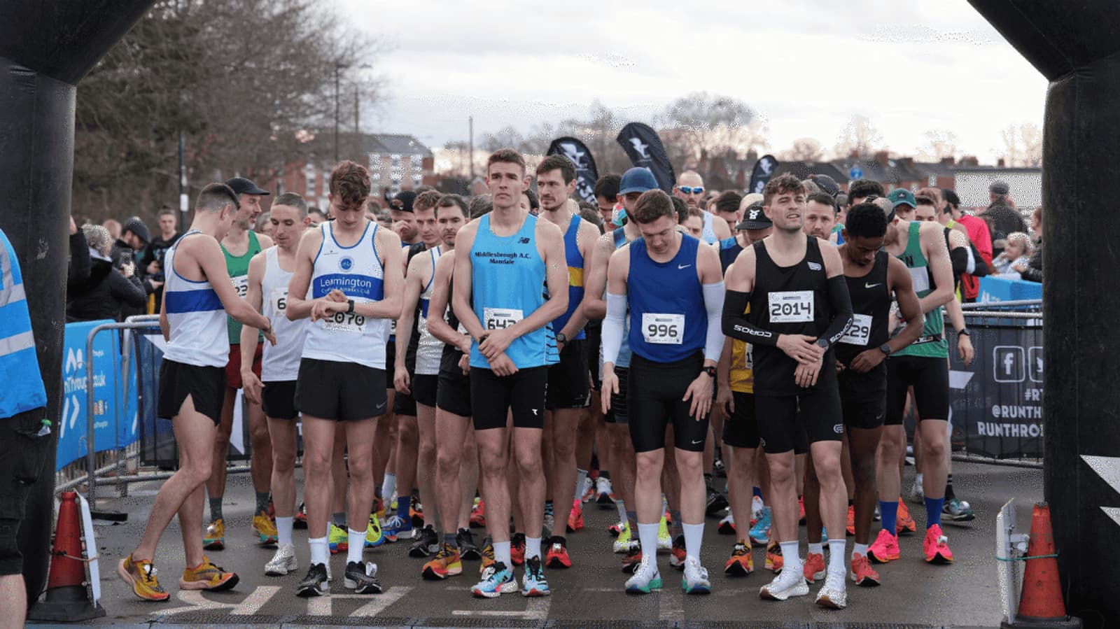 A group of male runners in athletic gear gather at the starting line of a race. They wear numbered bibs and colorful running shoes. The sky is overcast, and there are spectators and trees in the background.