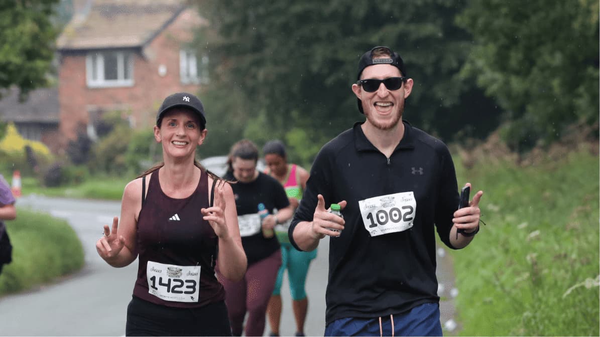 Two runners smile and gesture peace signs during a race, wearing numbered bibs 1423 and 1002. The woman wears a black cap and maroon tank top, and the man wears a black cap and shirt. Other runners are seen in the background on the road.