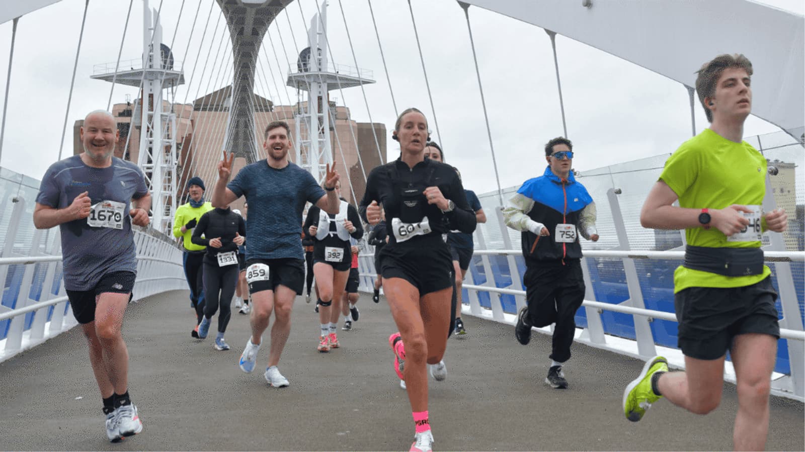 A group of runners crossing a modern suspension bridge during a race. They wear athletic gear and have race numbers on their shirts. The background shows part of the city skyline and an overcast sky.