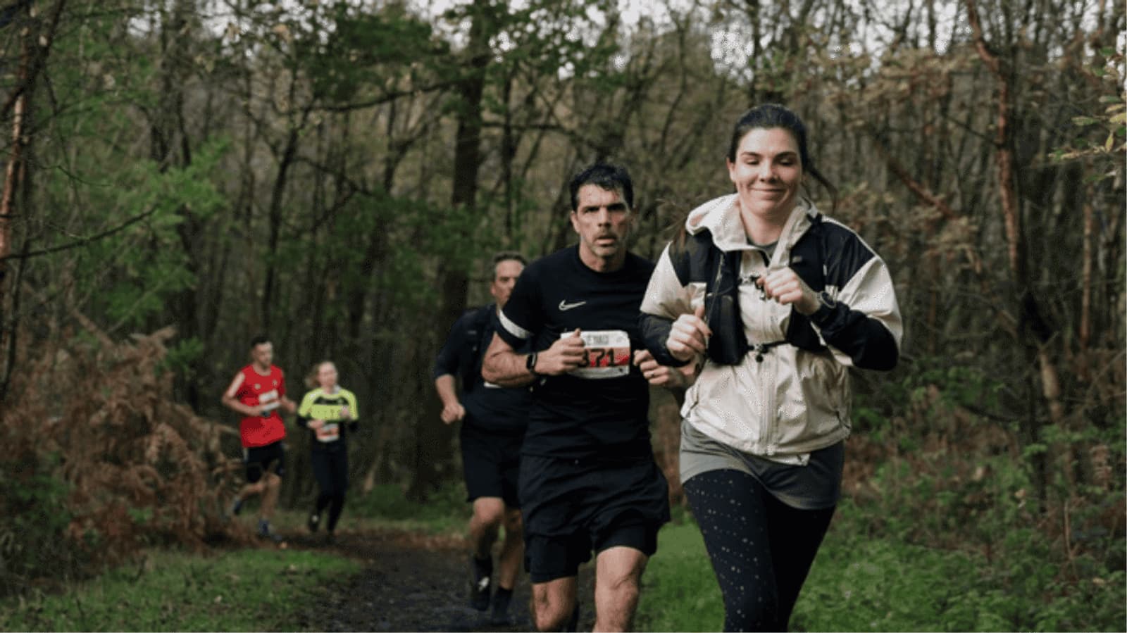 Several people are running on a wooded trail; a woman in a light jacket smiles in the foreground, while other runners follow behind her through the forest.