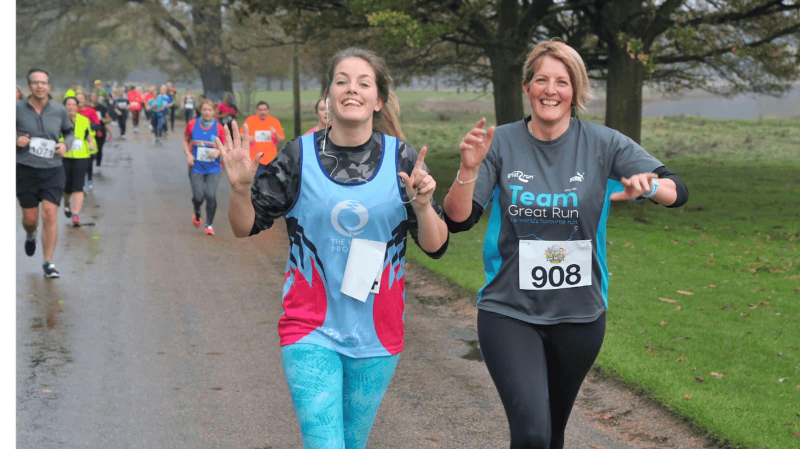 Two women are running in a race, smiling and making peace signs. They wear race bibs numbered 6 and 908. Other participants are visible in the background on a pathway flanked by grass and trees. The weather appears cool and overcast.