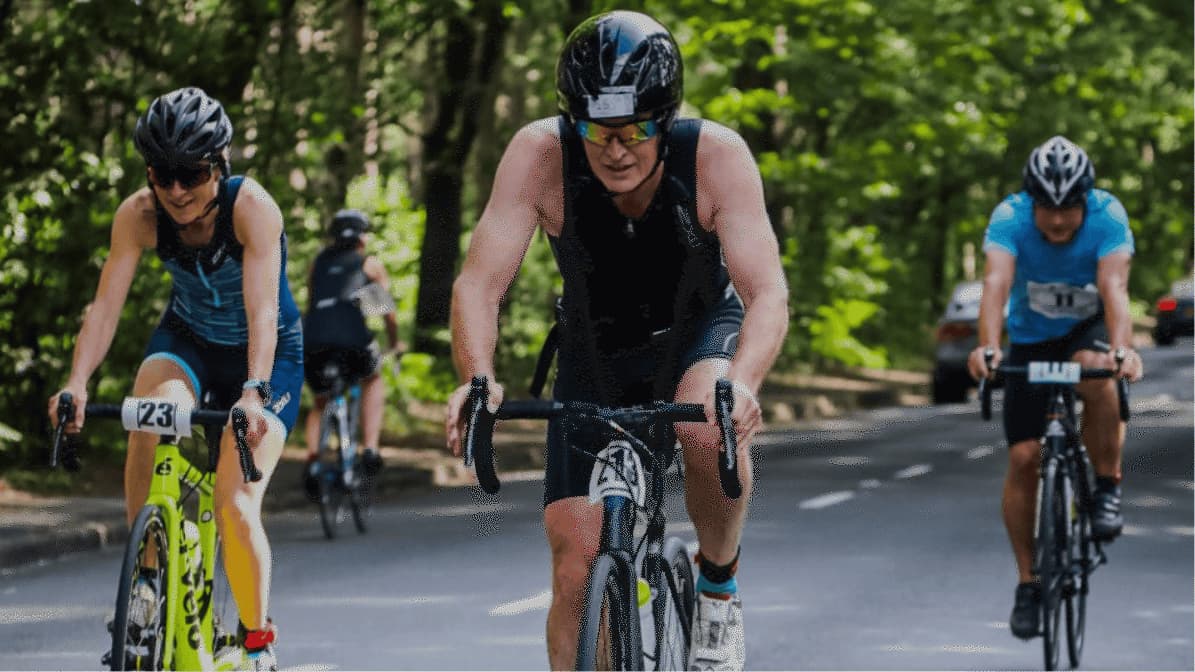 Three cyclists wearing helmets and athletic gear ride on a tree-lined road during a race. Two are in the foreground, one with a yellow bike, and one cyclist follows slightly behind. Race numbers are visible on their bikes.