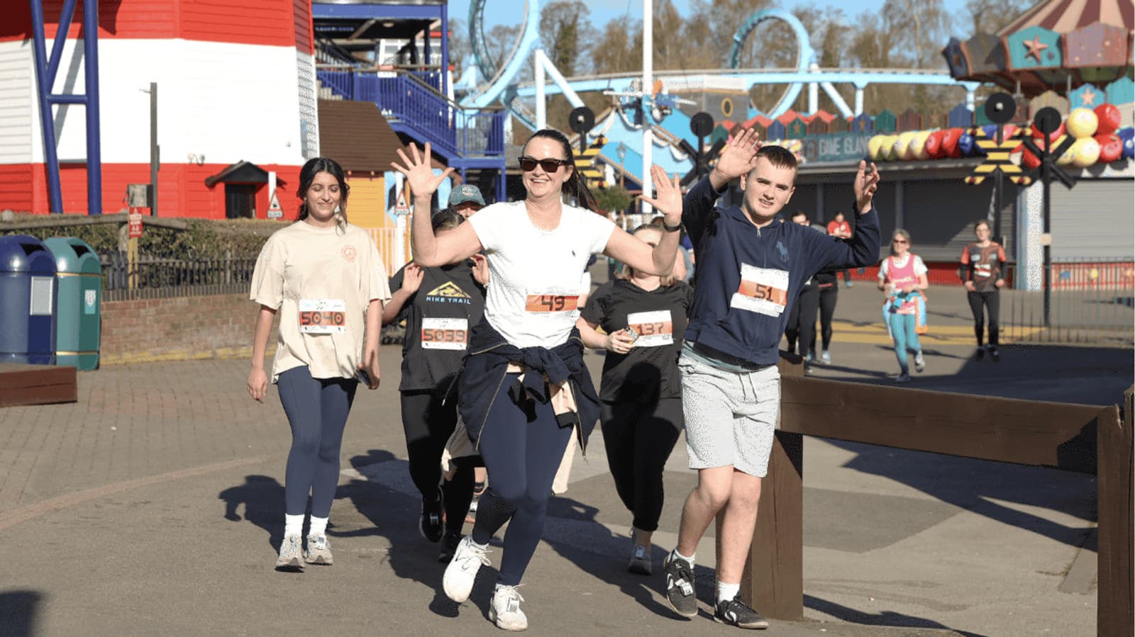 A group of people wearing numbered race bibs jog through an amusement park. One woman in front smiles and raises her arms in celebration, while others follow behind her. It is a sunny day, and colorful rides are visible in the background.