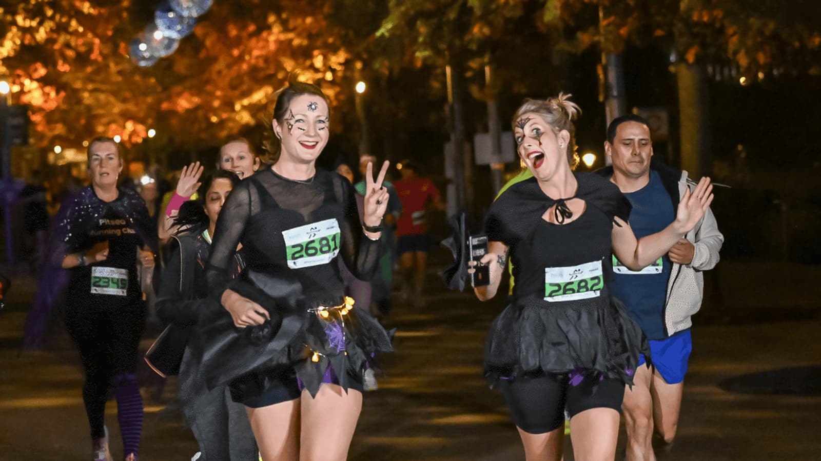 Participants in costume joyfully run a nighttime race. Two women in black outfits, one giving a peace sign, lead the cheerful group. Bib numbers and trees with lights are visible in the background.