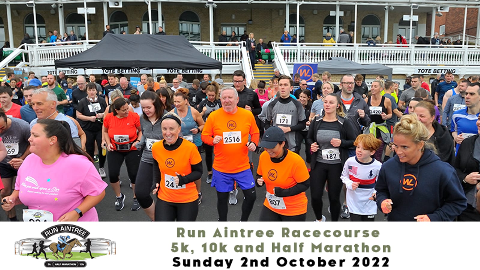 A large group of runners at the starting line of the Run Aintree Racecourse event on October 2, 2022. Participants of various ages and abilities are set to compete in 5k, 10k, and half marathon races. Spectators and the racecourse building are visible in the background.