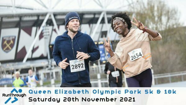 A man and a woman run together during the Queen Elizabeth Olympic Park 5k & 10k event on Saturday, 20th November 2021. Both are smiling and wearing race bibs (2892 and 2654). The West Ham United football stadium is visible in the background.
