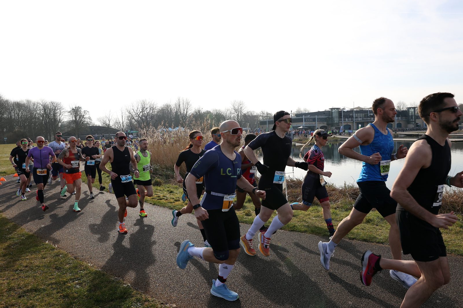 A group of runners, wearing athletic gear and race bibs, participate in a race on a paved path beside a body of water, with trees and buildings visible in the background.